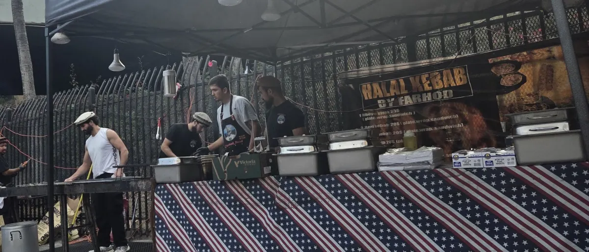 The stand under the concrete overpass, crew at the grill, flag-print tablecloth and the Halal Kebab by Farhod banner