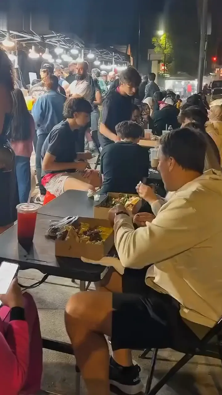 Guests eating from kraft boxes at the long table beside the stand at night